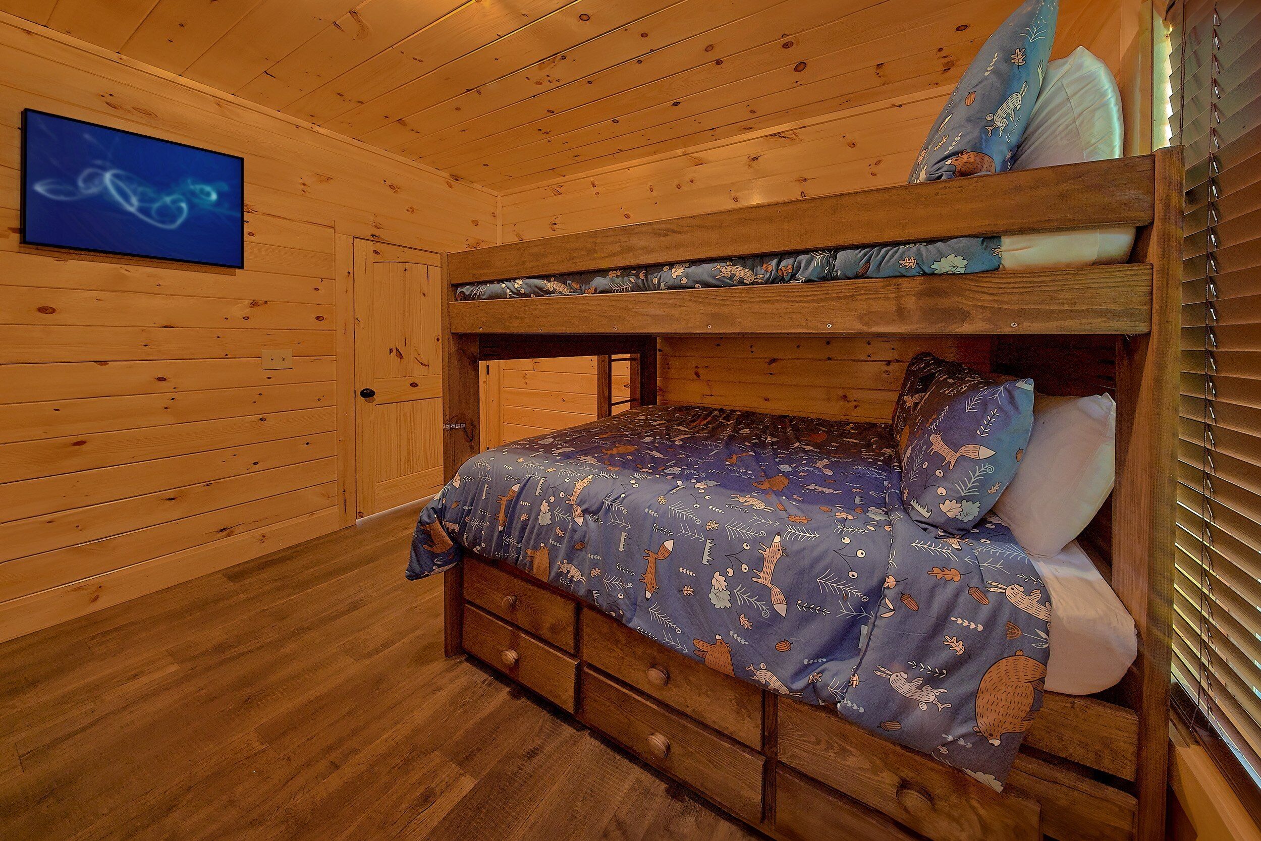 Ein Stockbett mit einem gemusterten Bettbezug und Kissen, Holzwände, ein an der Wand befestigter Fernseher und ein Fenster mit Jalousien.