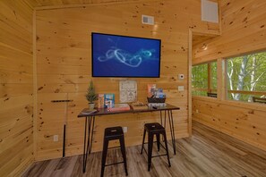 A wooden interior with a TV mounted on the wall, a small table with board games, two stools, and a window with a view of trees. - Misty Shadows Splash | Private Pool, Hot Tub, Game Room, Fireplace + Free Attraction Tickets (Wears Valley)