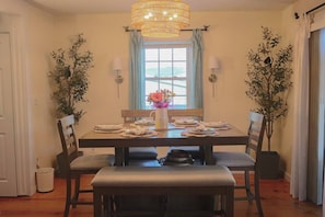 A dining area with a table set for four, flanked by two potted plants, a window with curtains, and a chandelier.