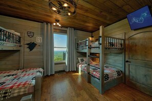 Eine Holzhütte mit Etagenbetten, Holzbalkendecke, einem Fenster mit Vorhängen und einem an der Wand befestigten Fernseher.