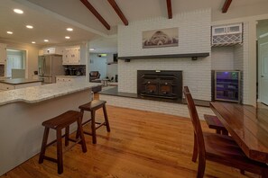 A modern kitchen with a white brick fireplace, wooden bar stools, and a dining area with a wooden table. - The Oasis | Sleeps 28, Private Pool, Theater, Mtn Views, Hot Tub, Fire Pit + Free Attraction Tickets (Sevierville)