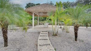 A covered outdoor seating area with a thatched roof, surrounded by palm trees and gravel ground. - Beautiful home with pool in Rancho Cartagena! (Cartagena)