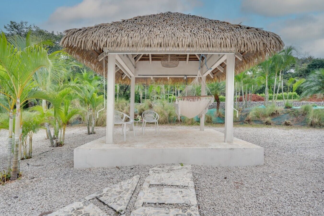 A thatched-roof gazebo with a white frame, surrounded by palm trees and a gravel path.
