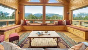 A cozy cabin interior with a large window seat overlooking mountains, wooden walls, and a central table. - 360 Mtn Views Close to Mt Princeton Hot Springs (Nathrop)