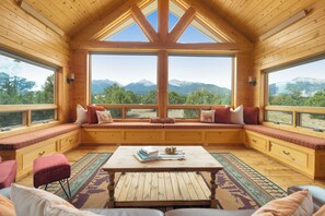 Ein gemütliches Cabin-Interieur mit einer großen Fensterbank mit Blick auf Berge, Holzwänden und einem zentralen Tisch.