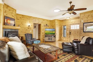 Interior acolhedor de uma cabana de madeira com lareira, móveis de couro, televisão e ventilador de teto.