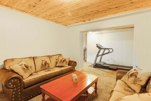 Sala de estar com sofá de couro marrom, mesa de centro de madeira e esteira ao fundo.