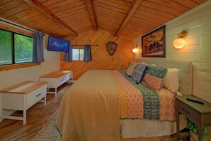 Ein Schlafzimmer mit Holzboden, einem Bett mit farbenfroher Decke, einem Schreibtisch, einem Stuhl und einem an der Wand befestigten Fernseher.