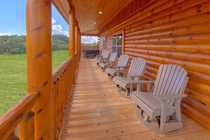 A wooden porch with Adirondack chairs and a table, overlooking a grassy field. - Livin' the Dream | Private Pool, Hot Tub, Game Room, Sleeps 12, Fireplace + Free Attraction Tickets (Pigeon Forge)