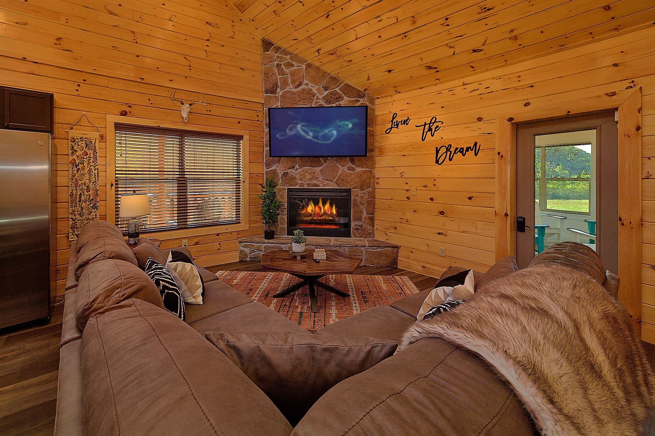 A cozy cabin interior with a stone fireplace, a television mounted above it, and a large window with blinds.