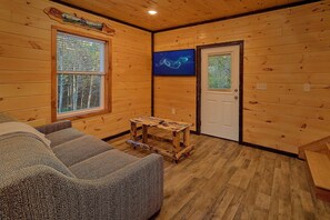 A log cabin interior with a wooden floor, a sofa, a small table, a door, and a window with a view of trees. - Relax II | Hot Tub, Game Room + Free Attraction Tickets (Sevierville)