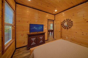 A wooden-paneled room with a bed, a television on a stand, and a door leading to another room. - Relax II | Hot Tub, Game Room + Free Attraction Tickets (Sevierville)