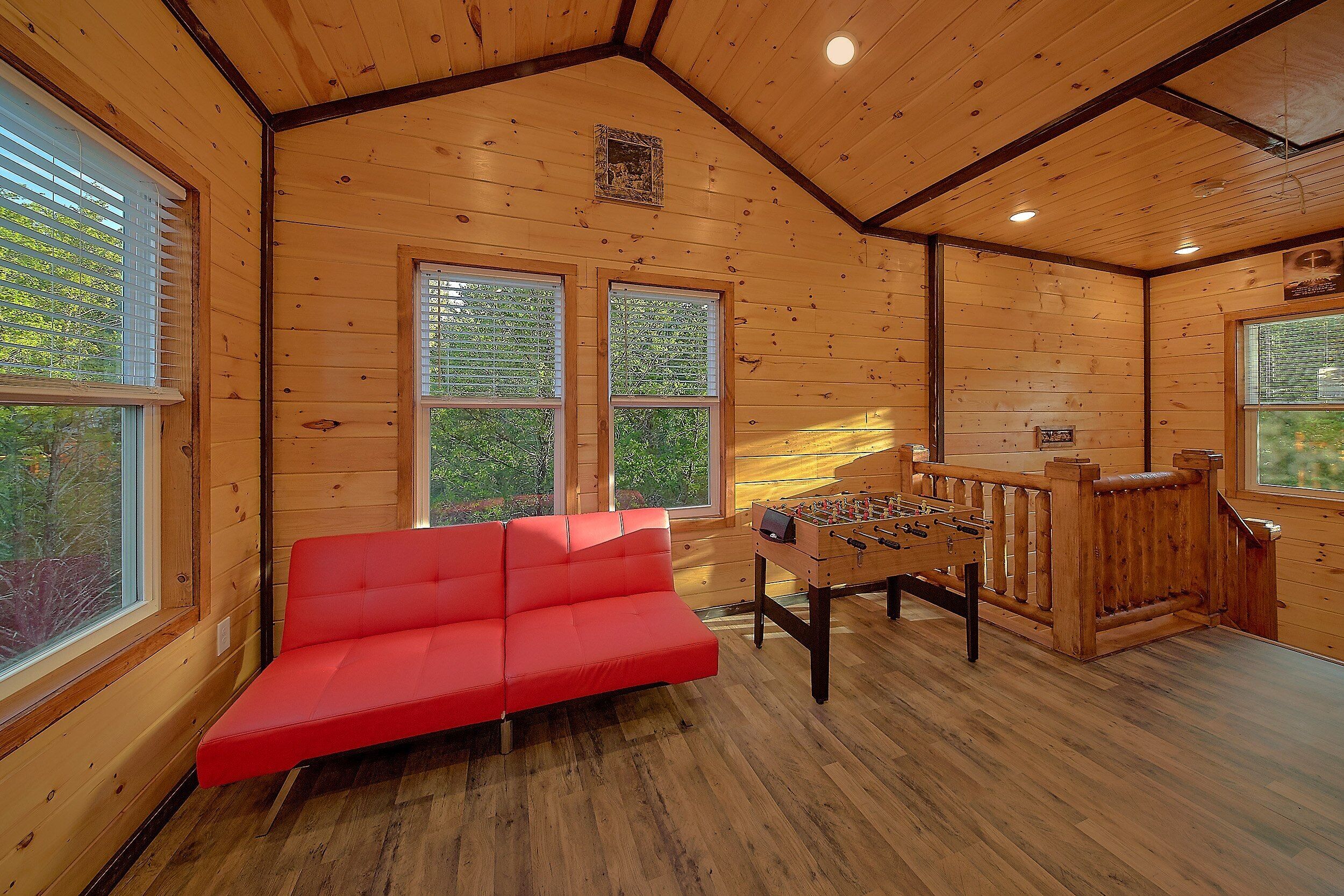 A cozy room with wooden walls, a red sofa, a foosball table, and large windows.