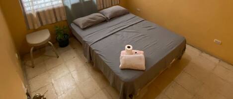 A small, single-bed room with a window, a bedside table, and a door.