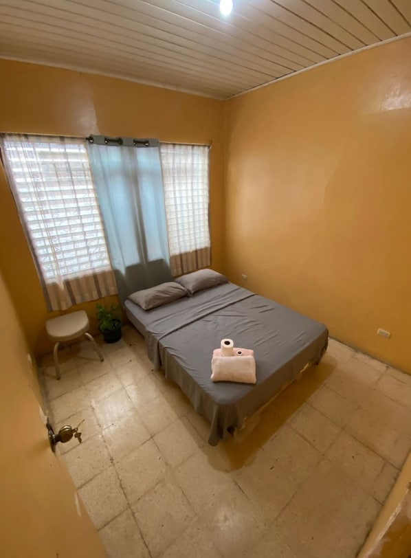 A small, single-bed room with a window, a bedside table, and a door.