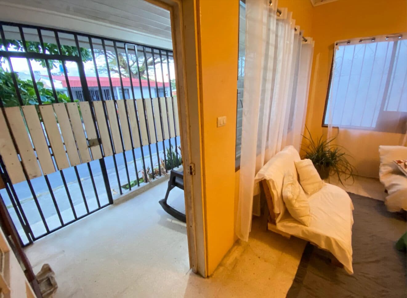 Habitació amb llit, finestra amb cortines, porta que dona a l'exterior i vista d'un carrer amb un cotxe aparcat.