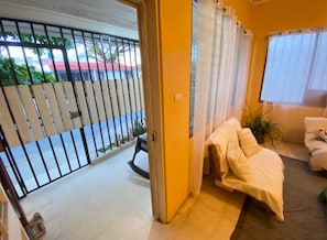 A room with a bed, a window with curtains, a door leading outside, and a view of a street with a parked car.