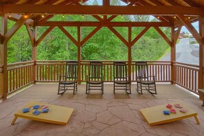 Un espace extérieur abrité, doté de chaises et de tables en bois, entouré d’une balustrade en bois, et offrant une vue sur des arbres verdoyants.