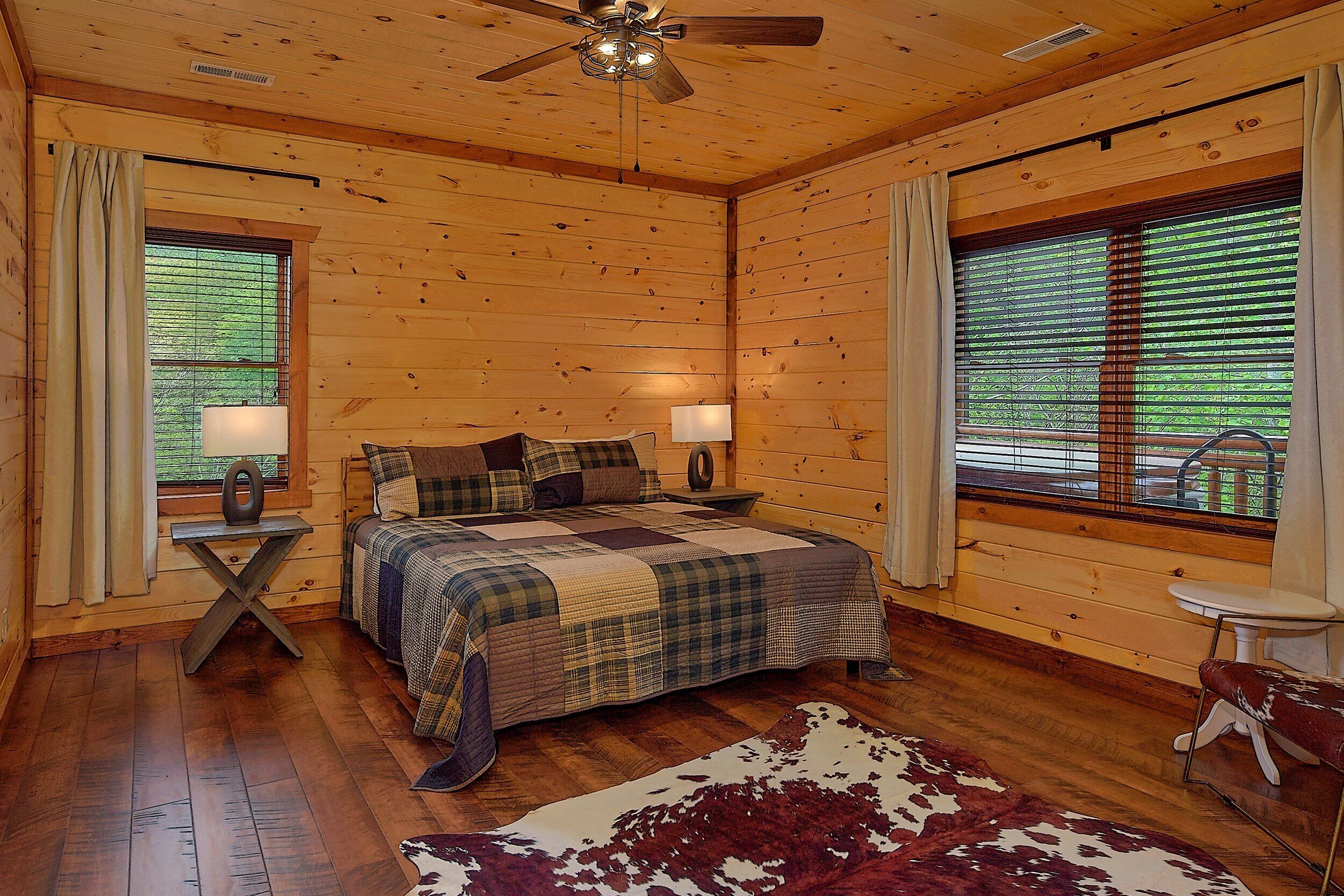 Ein Schlafzimmer in einer Blockhütte mit einem Bett, zwei Nachttischen, einem Deckenventilator und großen Fenstern mit Vorhängen.