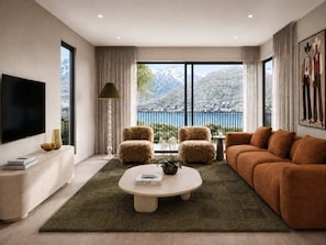 A modern living room with a large flat-screen TV, a sofa, a coffee table, and a view of mountains through large windows. - Lakehouse 63 – Brand- New Lakefront Villa with Spa (Queenstown)