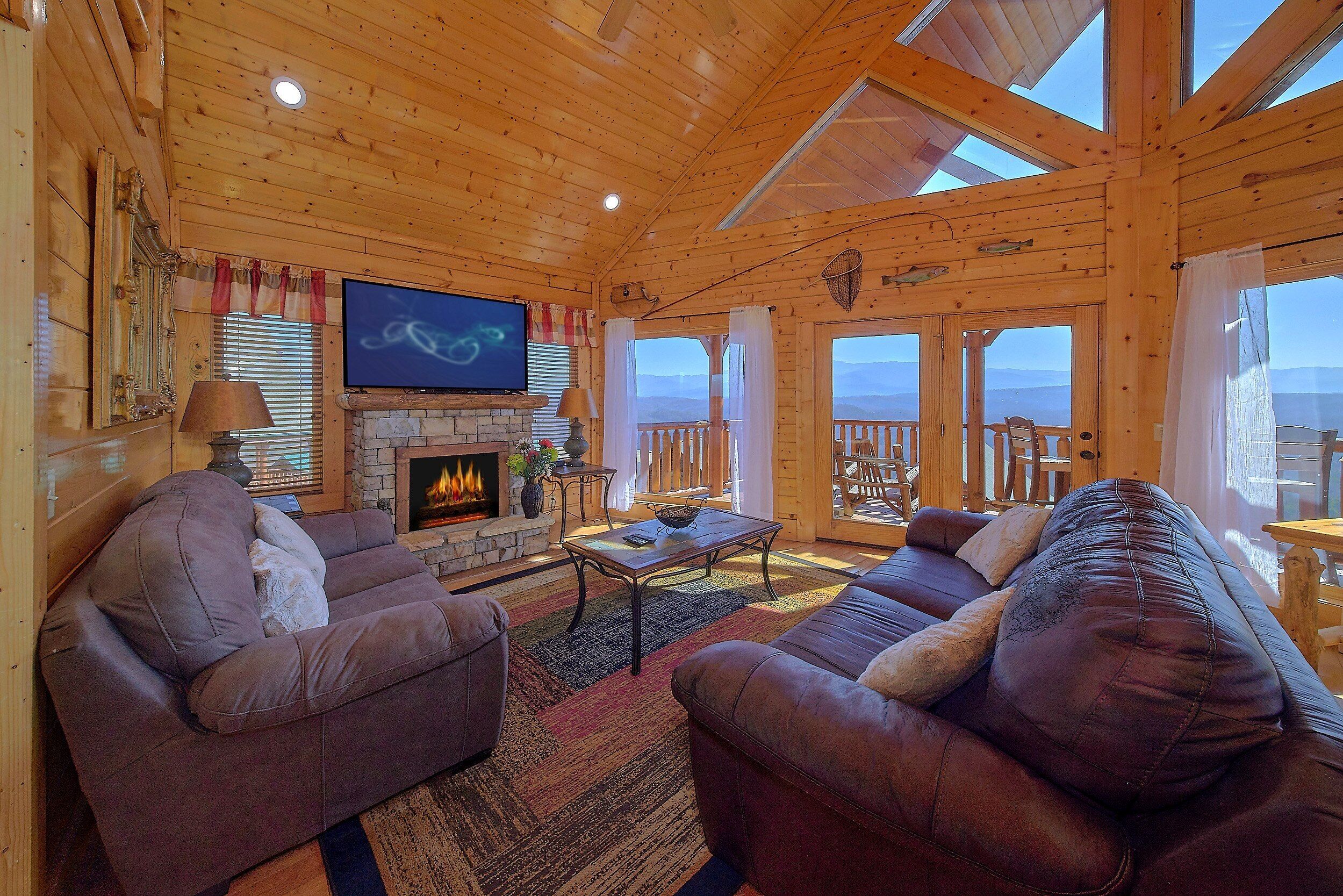 A log cabin interior with a fireplace, leather sofas, a television, and a balcony with a view.