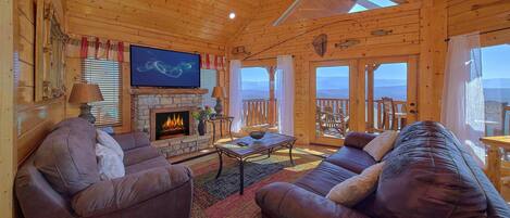 A log cabin interior with a fireplace, leather sofas, a television, and a balcony with a view.