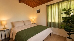Ein Schlafzimmer mit einem Bett, Nachttischlampen, einer Zimmerpflanze und einem Fenster mit Vorhängen.