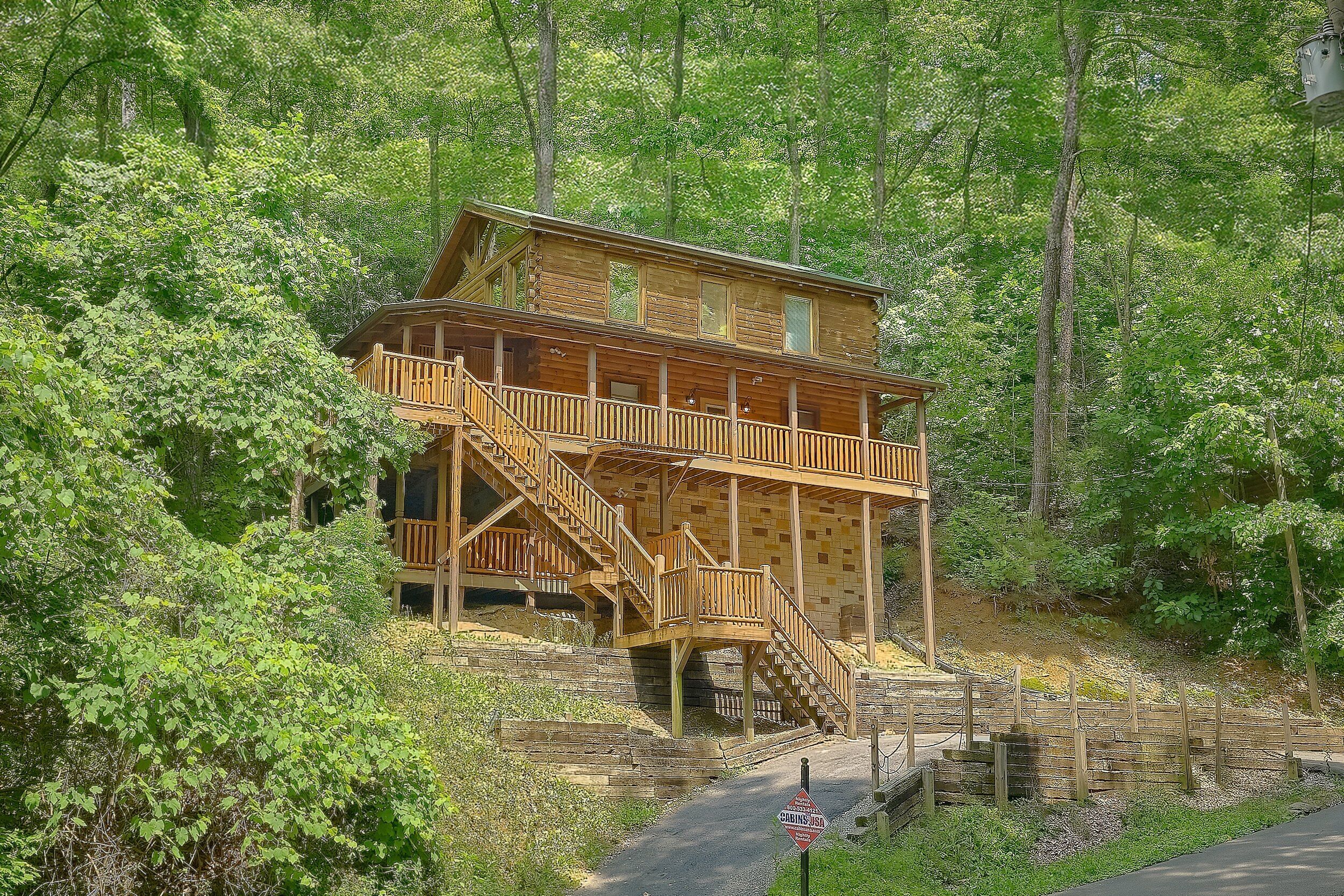A wooden cabin with a staircase leading to a balcony, surrounded by dense forest.