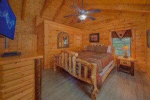 A wooden cabin bedroom with a bed, a ceiling fan, a television, and a window with a bear decoration. - Above the Rest | Game Room | Hot Tub | Fireplace (Sevierville)