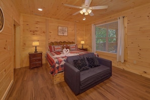 A wooden cabin bedroom with a bed, a sofa, a nightstand, a lamp, a ceiling fan, and a window with blinds. - Amazing Mountain Cabin Rental (Sevierville)