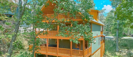 A two-story wooden cabin with a balcony and a wrap-around porch, surrounded by trees.