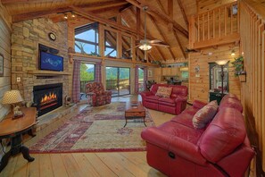 A spacious living room with a fireplace, red leather sofas, a television, and a large window overlooking a scenic outdoor area. - Timberwolf | Smoky Mtn Views, Hot Tub, Game Room, King Suites, Fireplace + Free Attraction Tickets (Wears Valley)