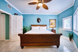 A bedroom with a wooden bed, a ceiling fan, a nightstand with a lamp, a door, and a window with blinds.
