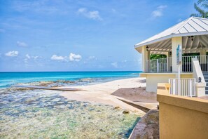 A beach with clear turquoise water, a sandy shore, and a building with a white roof and balcony.