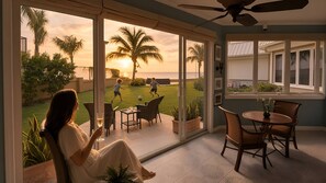 A room with a large sliding glass door leading to a patio with a view of palm trees and a sunset. Inside, there is a dining area with a table and chairs.