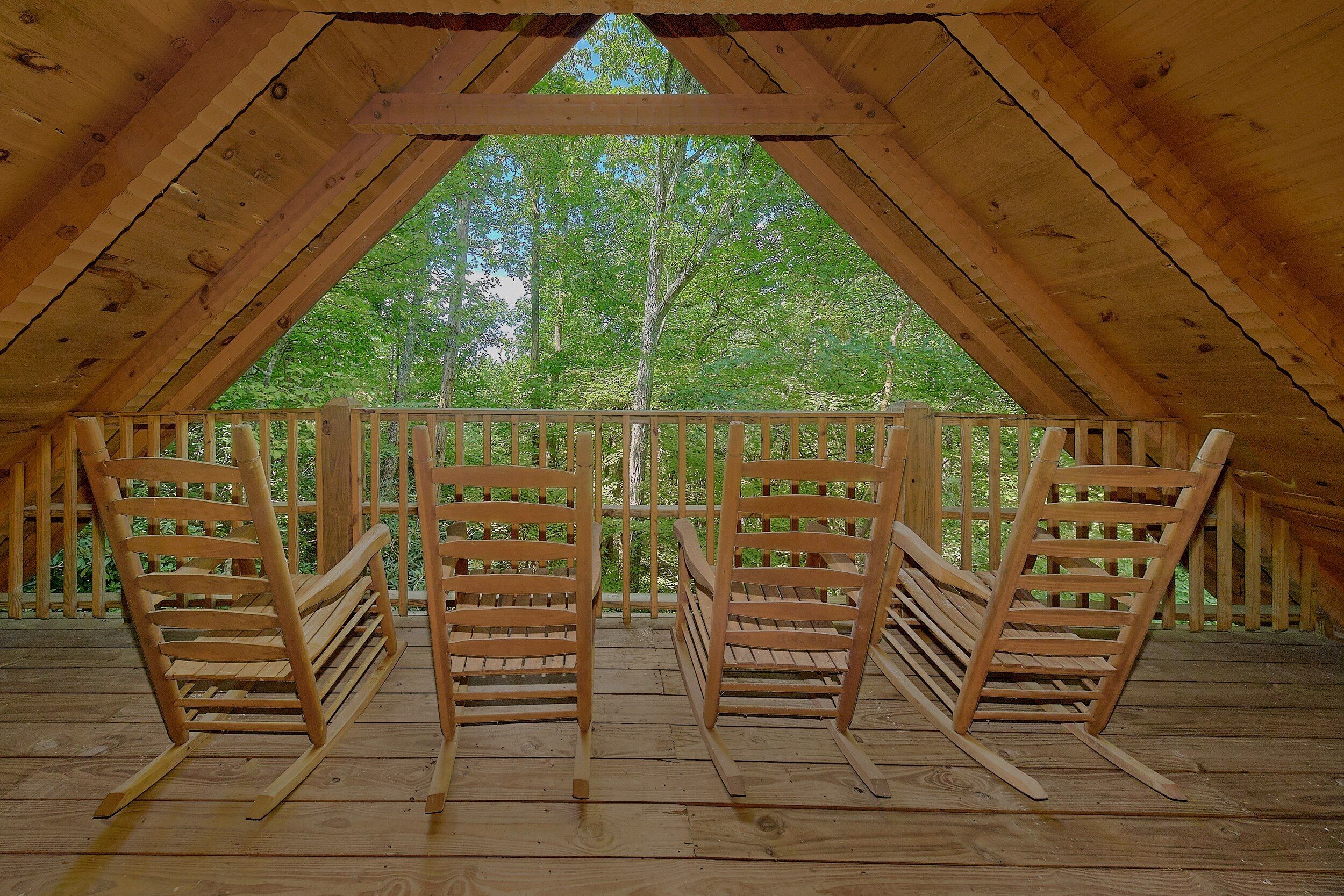 An attic room with a wooden floor and slanted ceiling, featuring three rocking chairs and a view of green trees.