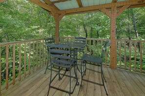A wooden deck with a metal table and chairs, surrounded by a wooden railing, set in a forested area. - Creek Song | Hot Tub, Fireplace + Free Attraction Tickets (Wears Valley)