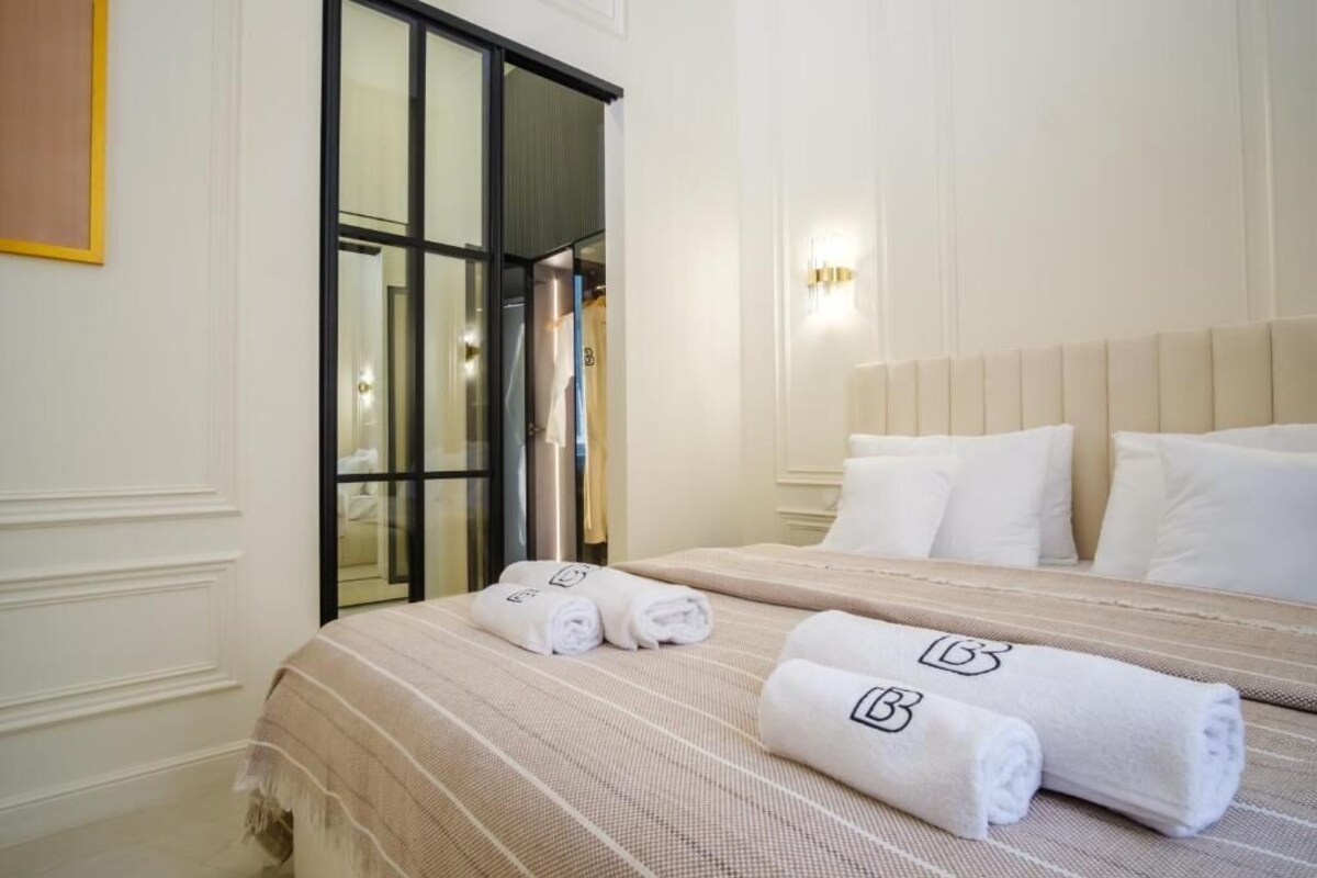 Ein Hotelzimmer mit einem großen Bett, zwei zusammengerollte Handtücher mit einem Logo, einem Fenster mit Jalousien und einer an der Wand montierten Leuchte.