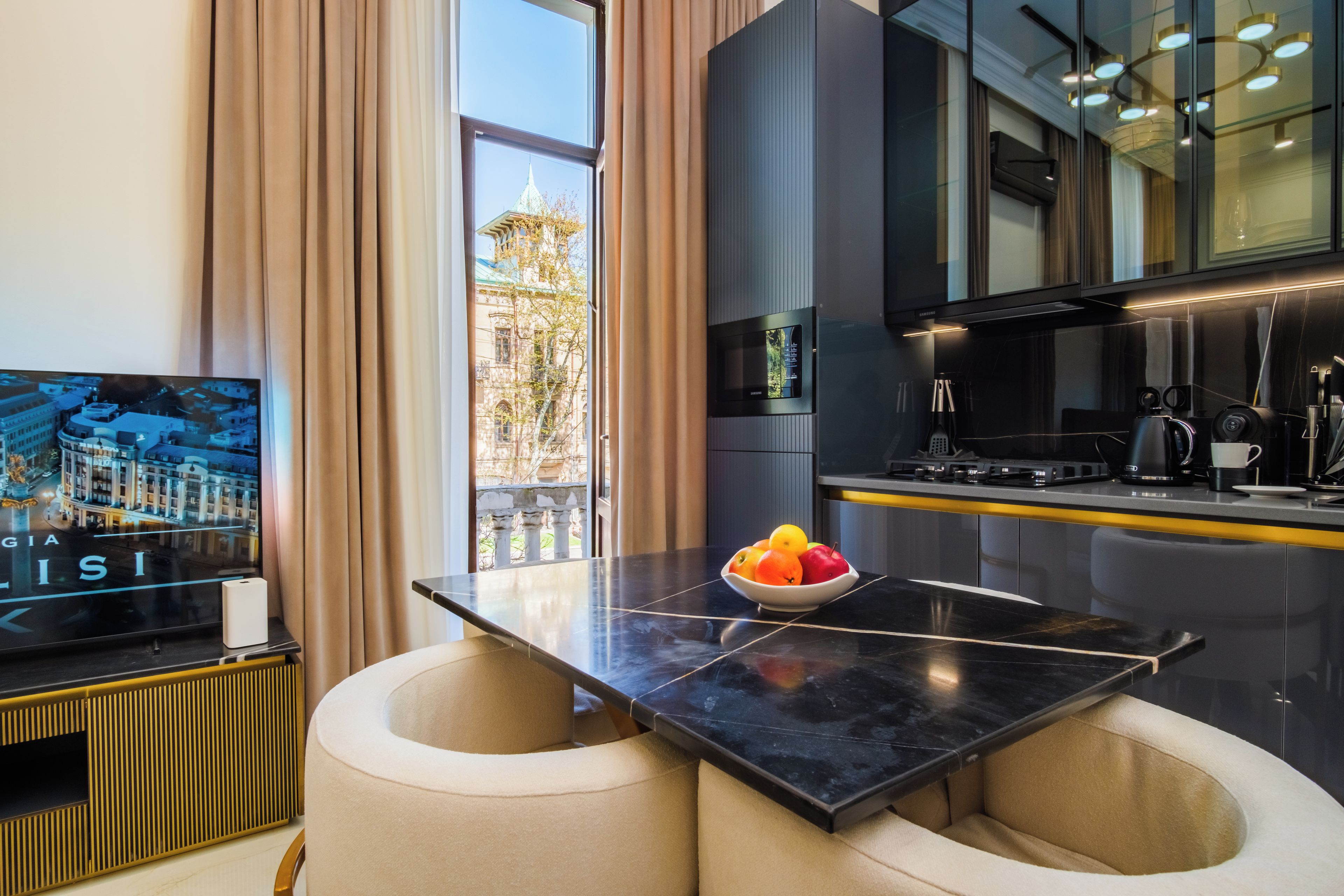 A modern kitchen with a black countertop, a bowl of fruit, and a television displaying a cityscape.