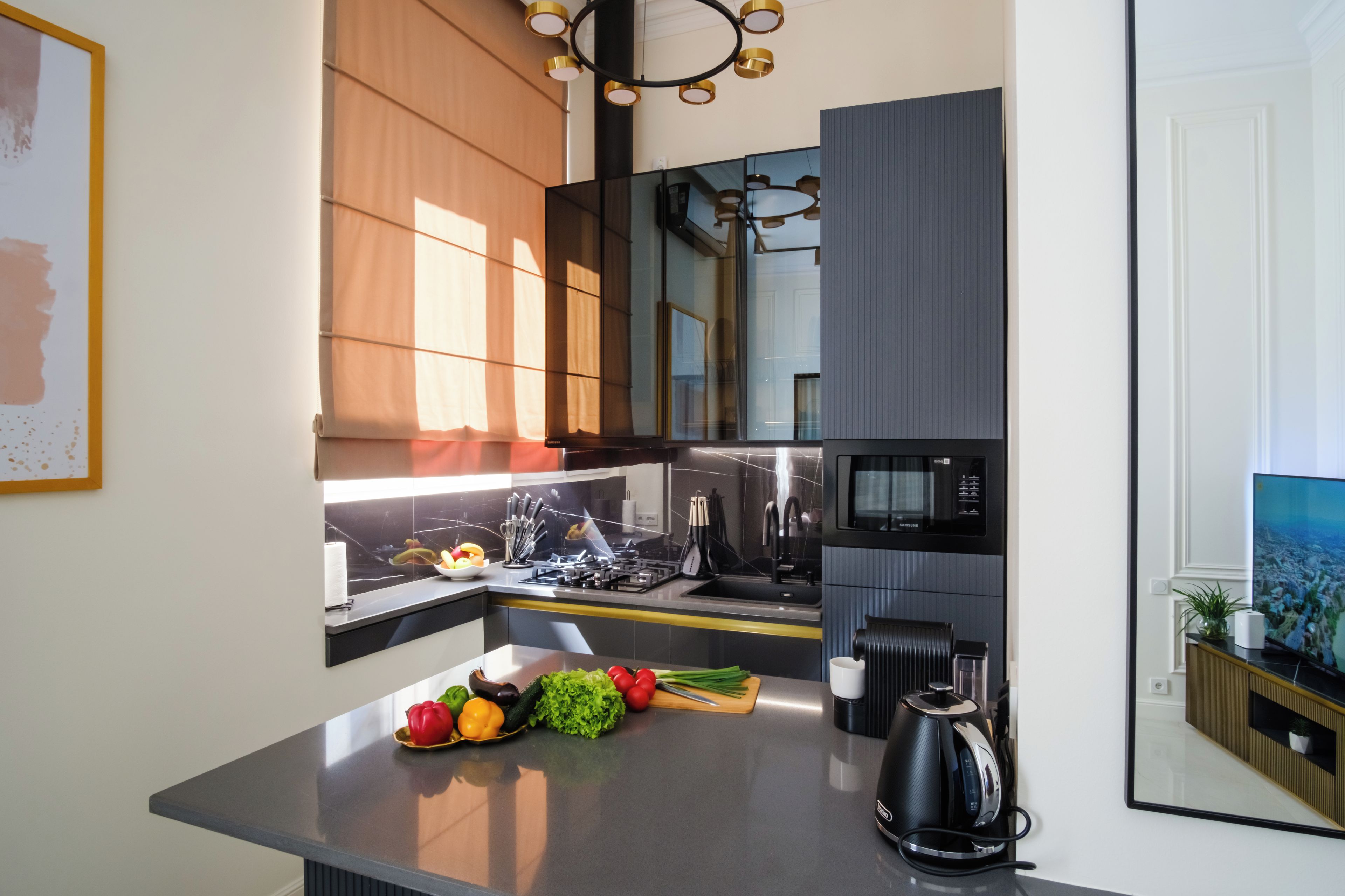 A modern kitchen with a built-in microwave, stove, and sink. There is a fruit bowl with various fruits and vegetables on the counter.