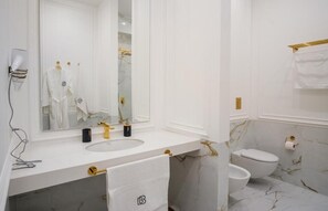 Ein Badezimmer mit einem weißen Waschtisch, einem großen Spiegel, einer goldenen Armatur, einer Toilette und Marmorwänden.