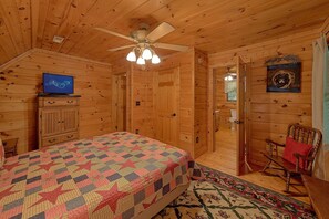A log cabin bedroom with a bed, a television, a ceiling fan, and a wooden door. - Moonlight Magic | Hot Tub, Resort Pool, Fireplace + Free Attraction Tickets (Wears Valley)