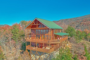 A wooden cabin with a balcony and a green roof, surrounded by trees and hills. - Lookout Lodge | Smoky Mtn Views, Hot Tub, Game Room, Sleeps 12, Fireplace + Free Attraction Tickets (Wears Valley)