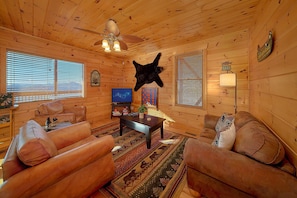 A cabin interior with a ceiling fan, a mounted bear head, wooden walls, a television, and a fireplace. - Lookout Lodge | Smoky Mtn Views, Hot Tub, Game Room, Sleeps 12, Fireplace + Free Attraction Tickets (Wears Valley)