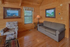 Un acogedor interior de cabaña con suelo de madera, un sofá gris, un sillón estampado, una mesita y dos cuadros en las paredes.