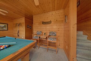 Interior de una cabaña de madera con mesa de billar, dos sillones, una mesita y una escalera.
