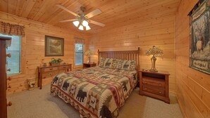 Une chambre à coucher chaleureuse, aménagée dans le style d’un chalet, avec un plafond en bois, un lit recouvert d’une courtepointe à motifs, deux tables de chevet avec des lampes et une fenêtre avec des stores.