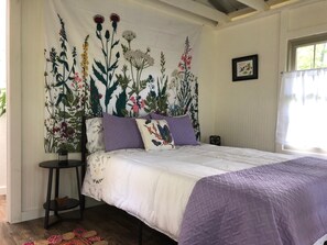 Een slaapkamer met een bed, een nachtkastje, een wandtapijt met bloemenmotieven en een raam met gordijnen.