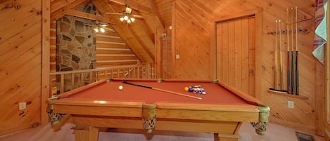 A wooden interior with a pool table, a ceiling fan, and a staircase leading to a stone wall.