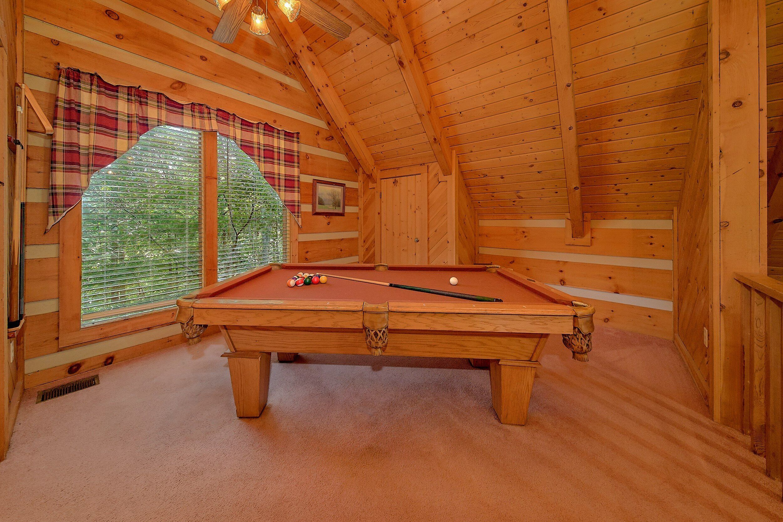 A wooden interior with a pool table, plaid curtains, and a window with blinds.